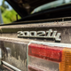 a close-up photo of the 2002tii badge on the back of a very dirty maroon bmw coupe from 1974